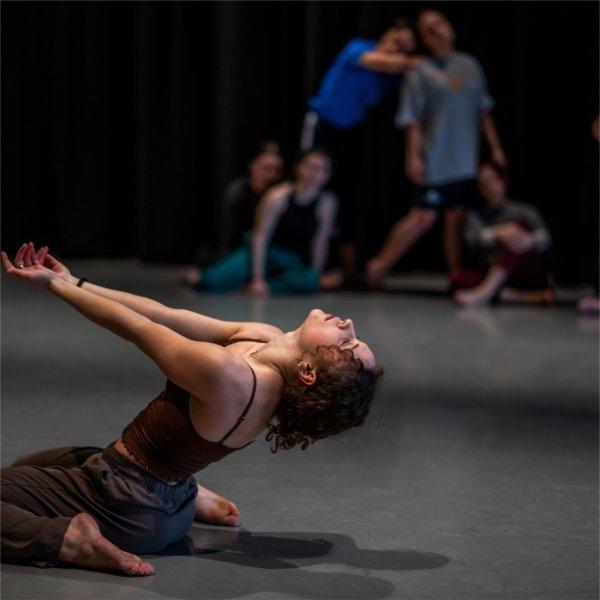 A dancer is on their knees, bending back with arms reaching out. Other dancers are in the background.